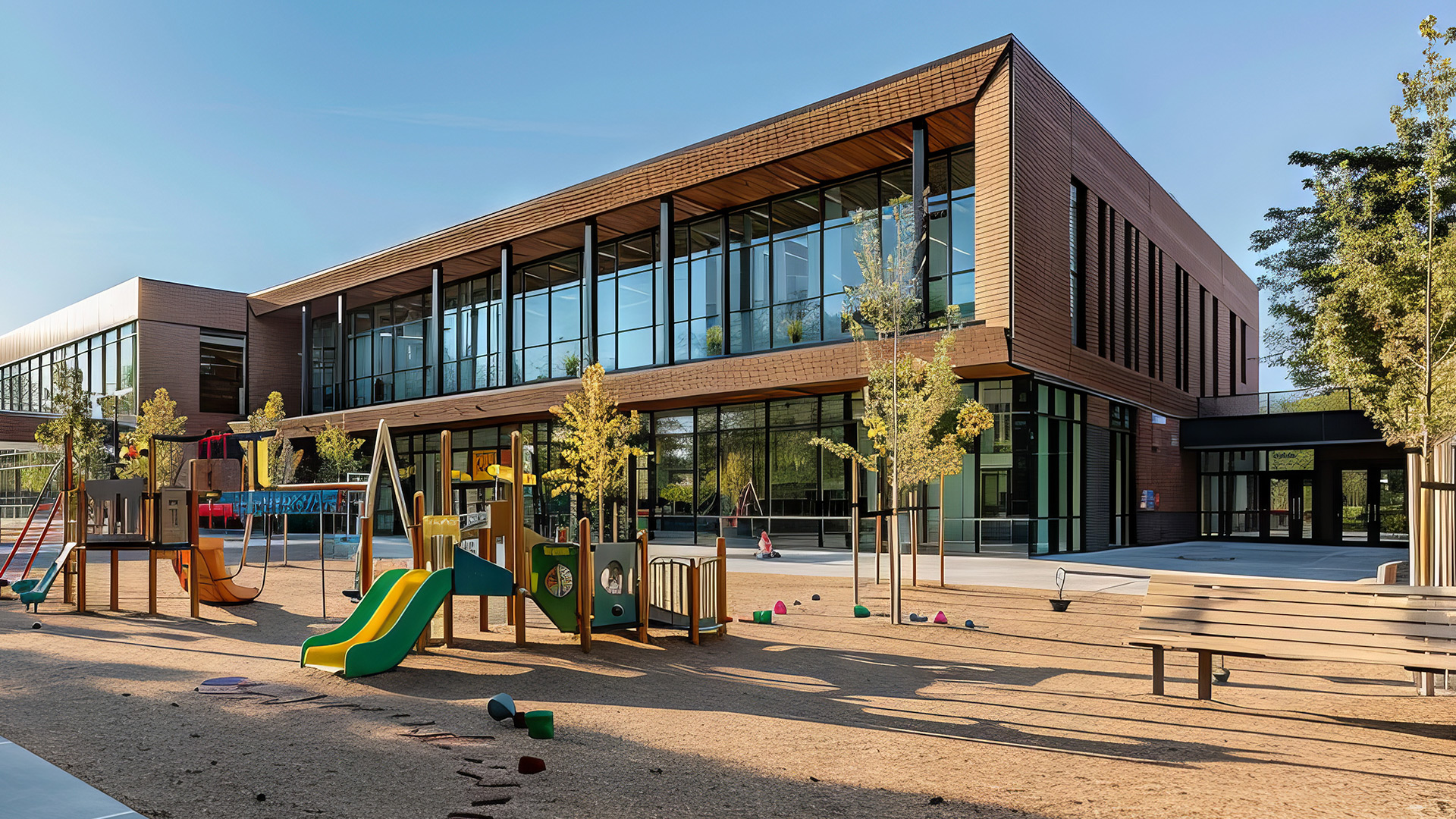 A modern two-story building with brown paneling and large glass windows stands behind a colorful playground with slides and a wooden park bench.