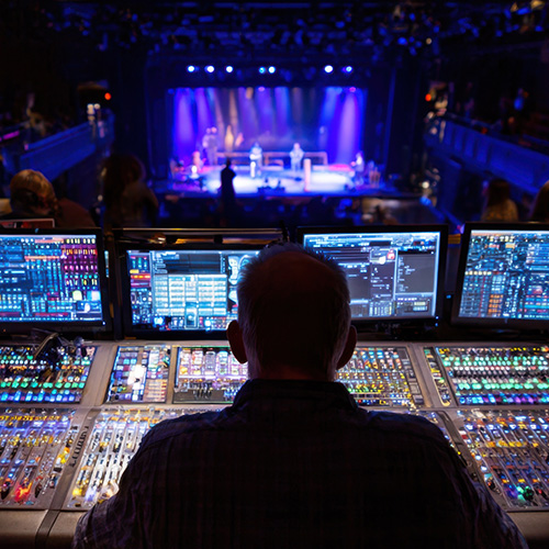 Live audio mixing console control room with multiple screens and blue stage lighting in commercial venue